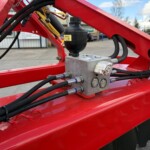 Close-up of a hydraulic control block mounted on a red metal frame, with a black reservoir and multiple hoses.
