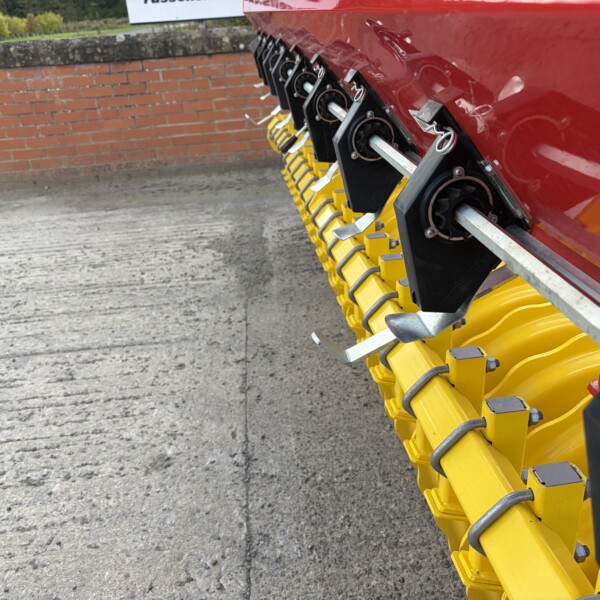 Close-up of yellow tines mounted on a red agricultural machine, brick wall in the background