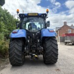 Rear view of a blue tractor with oversized rear tires, parked in a driveway with a green hedge on the left and brick houses in the background.