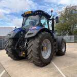 Front-and-side view of a blue agricultural tractor with oversized rear tires parked in an outdoor lot, fence and trees in the background.