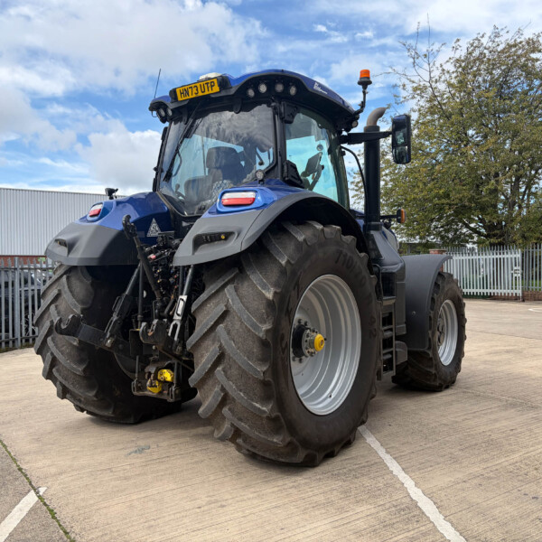 Front-and-side view of a blue agricultural tractor with oversized rear tires parked in an outdoor lot, fence and trees in the background.
