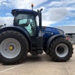 Blue New Holland tractor photographed from the front-left, showing oversized rear tires and orange roof beacon in a parking lot.