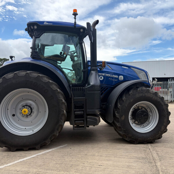 Blue New Holland tractor photographed from the front-left, showing oversized rear tires and orange roof beacon in a parking lot.