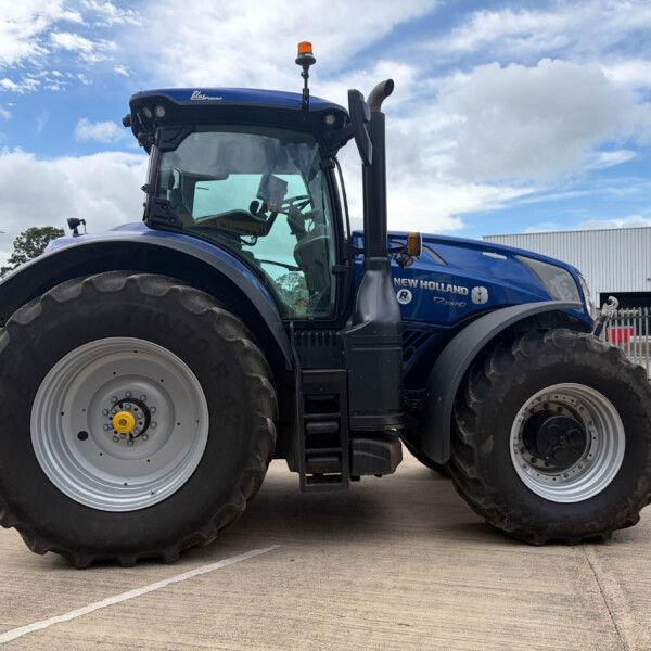 Front-left view of a blue New Holland tractor with large tires and an orange beacon, parked in an outdoor lot behind a fence.