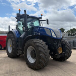 Blue New Holland tractor with oversized front tires and orange roof beacons parked in a lot against a cloudy sky and fence in the background.