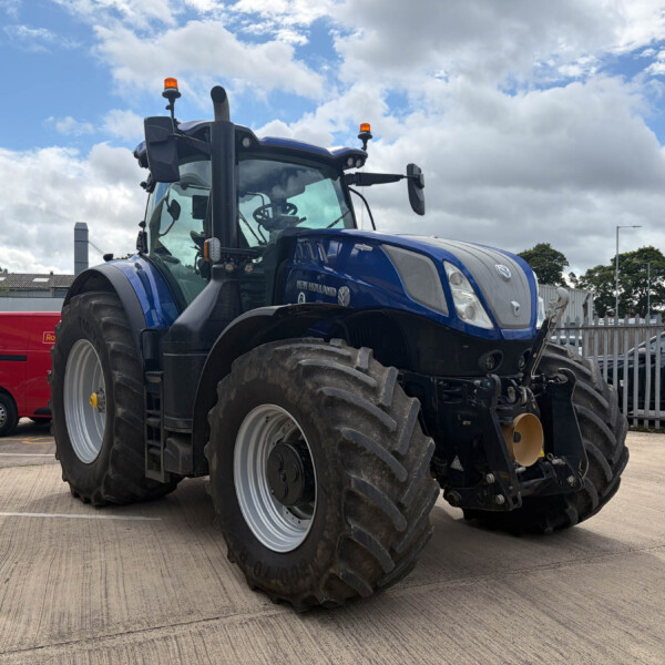 Blue New Holland tractor with oversized front tires and orange roof beacons parked in a lot against a cloudy sky and fence in the background.