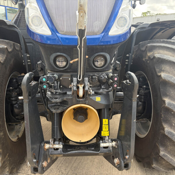 Front view of a blue tractor with oversized muddy tires, showing hydraulic linkages, central hitch, and tool attachments.