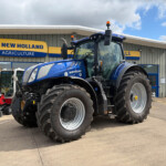 Blue New Holland tractor parked outside an agricultural dealership with a yellow-and-blue New Holland sign in the background, large tires visible in the foreground.
