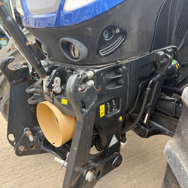 Close-up of a blue-fronted excavator with black metal frame, hydraulic linkages, and large dusty tires visible in a construction yard.