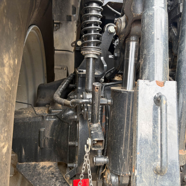 Underside of a dusty vehicle showing a coil spring, shock absorber, and various metal linkages and hoses in a cramped chassis area.