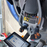 Construction vehicle cab with color-coded switches, a joystick, and a tablet showing controls in use.