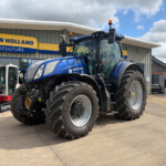 Blue New Holland Agriculture tractor parked in front of dealership sign in a light-industrial lot.