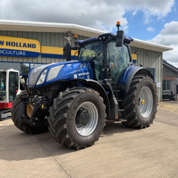 Blue New Holland Agriculture tractor parked in front of dealership sign in a light-industrial lot.