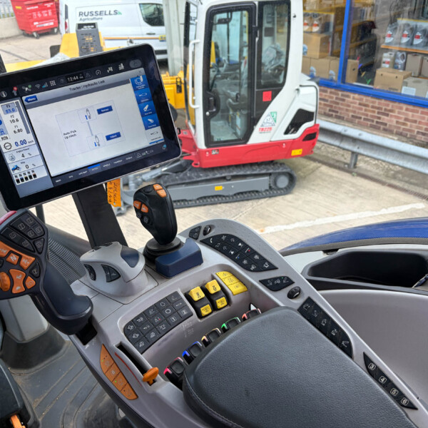 Construction vehicle cockpit interior with touchscreen display and multiple control joysticks/levers visible. Outside: a compact excavator and workshop window.