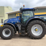 Blue New Holland tractor viewed from the front in a dealership lot, large tires and cab visible against a yellow-and-blue sign building.