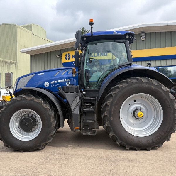 Blue New Holland tractor viewed from the front in a dealership lot, large tires and cab visible against a yellow-and-blue sign building.