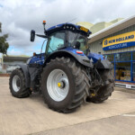 Blue New Holland tractor parked outside an agriculture store with a bright yellow and blue sign behind it.