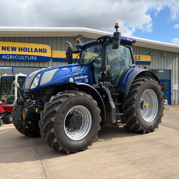 Blue New Holland tractor parked outside an agricultural dealership with a yellow-and-blue New Holland sign in the background, large tires visible in the foreground.