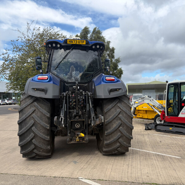 Rear view of a blue agricultural tractor with oversized tires, parked in an industrial yard with other equipment nearby.