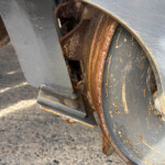 Close-up of a rusted metal wheel and brackets on industrial equipment, with chipped blue-gray paint and dirt nearby.
