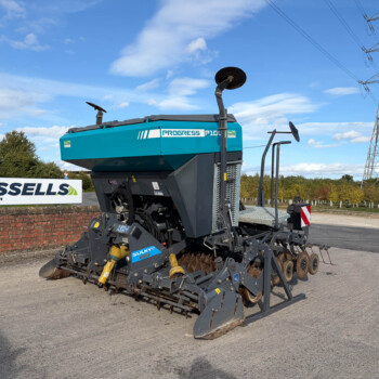 Blue and gray road construction machine with a large rotating cutter wheel sits on a paved lot, Russell's sign in the background, power lines visible behind.