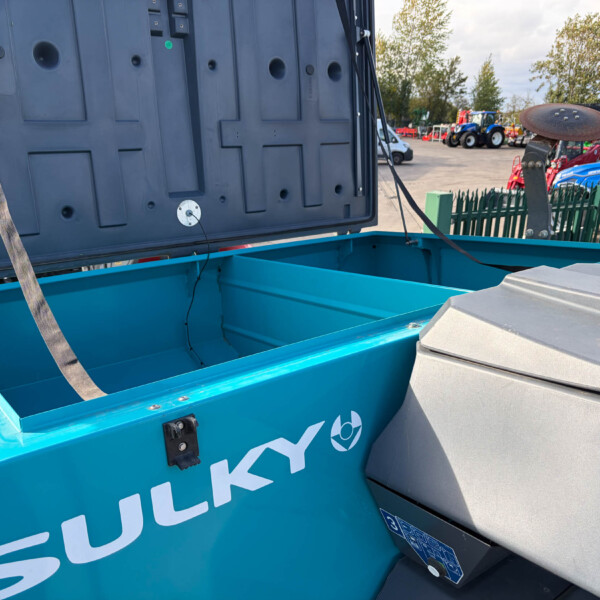 Blue open-lid dumpster in foreground with gray lid, straps, and empty interior, at an outdoor event with tractors in the background.