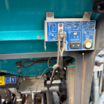 Close-up of a teal industrial machine showing a blue labeled control panel with levers and metal clamps, and various hoses nearby.