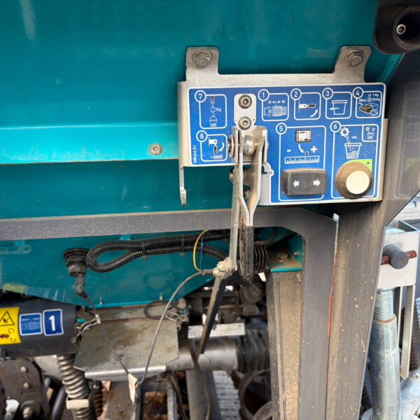 Close-up of a teal industrial machine showing a blue labeled control panel with levers and metal clamps, and various hoses nearby.