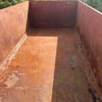 Interior of an empty, rust-coated metal dumpster outdoors, with peeling paint and a shadow at the bottom reading light and shade across the floor.