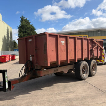 Red dump trailer parked in a paved lot beside a beige industrial building, with a forklift in the foreground and red waste bins along a fence