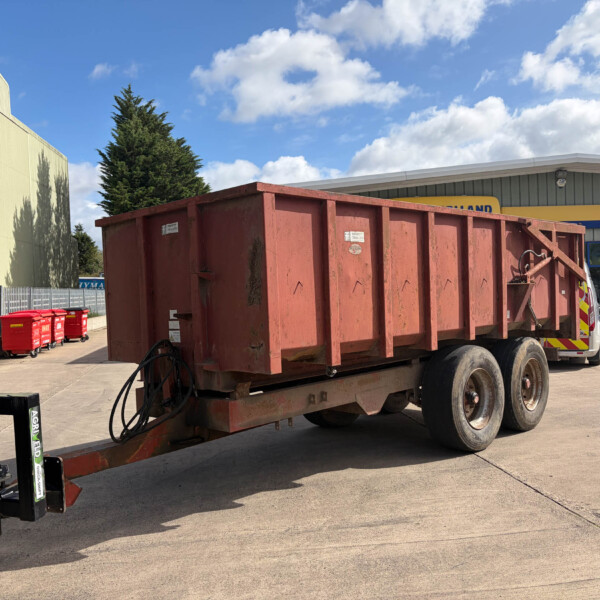 Red dump trailer parked in a paved lot beside a beige industrial building, with a forklift in the foreground and red waste bins along a fence