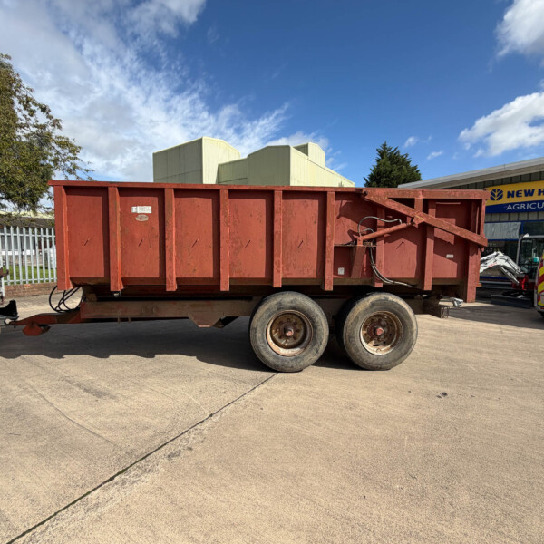 Red, rusted dump trailer parked in a lot with a New Holland Agriculture building in the background on a sunny day.