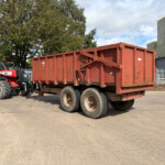 Red tractor with a large red dump trailer in an industrial yard, with a green warehouse and trees in the background