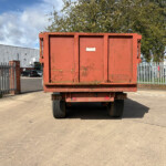 Red, weathered dumpster on a flatbed trailer in an industrial lot with a metal fence and brick building nearby.