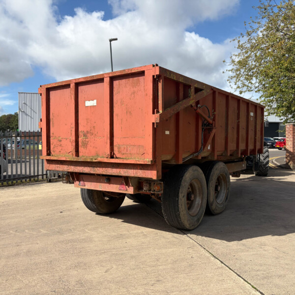 Red/orange dump truck trailer parked in an open lot with a metal fence and leafy trees nearby on a sunny day.