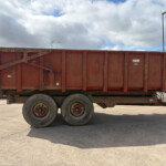 Rusty red industrial dump trailer on a multi-axle trailer in a paved yard, with a fence and a building in the background.