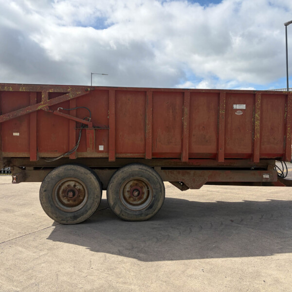 Rusty red industrial dump trailer on a multi-axle trailer in a paved yard, with a fence and a building in the background.