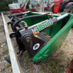 Close-up of a green industrial skip with hydraulic hoses and metal components; red tractors and other machinery are in the background.