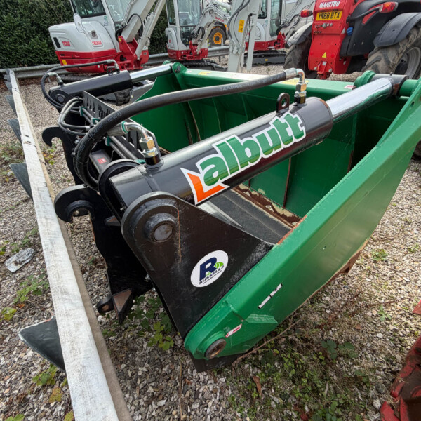 Close-up of a green industrial skip with hydraulic hoses and metal components; red tractors and other machinery are in the background.