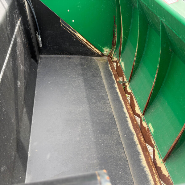 Interior view of a green dumpster with a black floor, showing rust along a seam and a small weed growing by the edge.