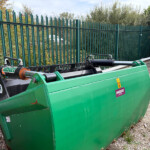 Green industrial waste container with a long hopper, labeled Hardox, parked beside a green metal fence on a gravel surface.