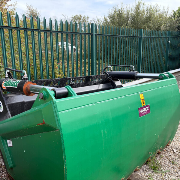 Green industrial waste container with a long hopper, labeled Hardox, parked beside a green metal fence on a gravel surface.