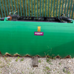 Green industrial container with hydraulic arms and a warning sticker, sitting on a gravel surface; labeled HARDOX wear plate on the side.
