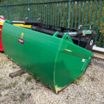 Green agricultural/industrial attachment with hydraulic cylinders, adjacent to a red machine on a gravel surface by a green fence.