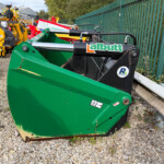 Green agricultural loader attachment with hydraulic cylinders parked on a gravel lot beside a green metal fence and other farming equipment in the background.