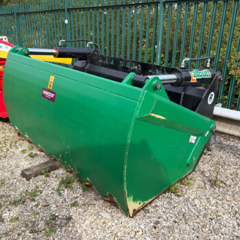 Green agricultural/industrial attachment with hydraulic cylinders, adjacent to a red machine on a gravel surface by a green fence.
