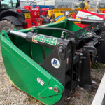 Green agricultural implement with hydraulic cylinders and hoses in a yard of tractors and farming equipment; gravel ground and wooden beam nearby