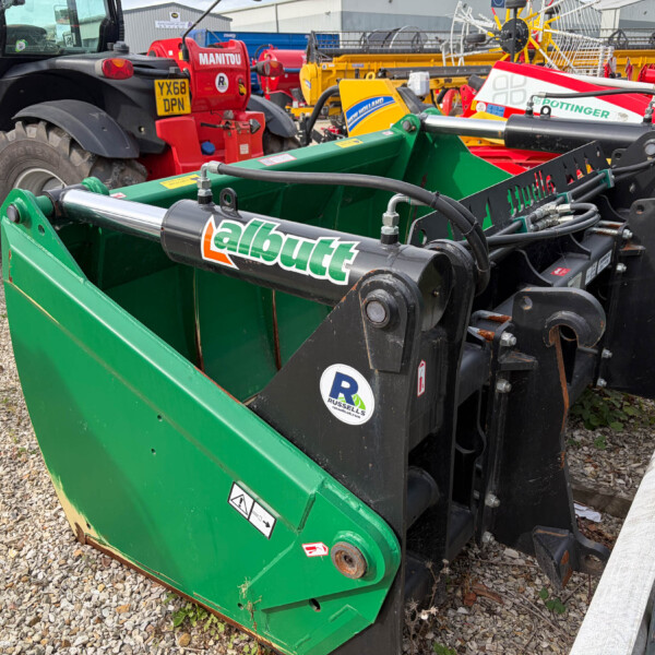 Green agricultural implement with hydraulic cylinders and hoses in a yard of tractors and farming equipment; gravel ground and wooden beam nearby
