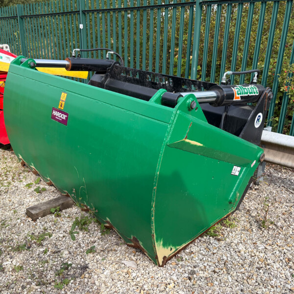 Green agricultural/industrial attachment with hydraulic cylinders, adjacent to a red machine on a gravel surface by a green fence.