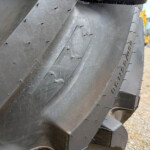 Close-up of a large black rubber track with stitched seams and rivets on a heavy construction machine; blurred yellow equipment visible in the background.